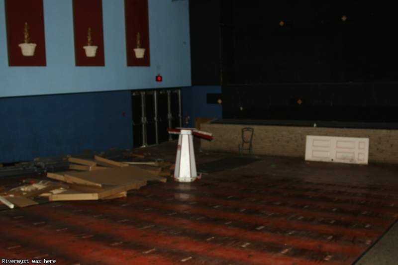 An emptied auditorium with blue walls and red panels of potted-plant artwork, its seats removed, wood scraps and an old door left on the floor in front of the stage.