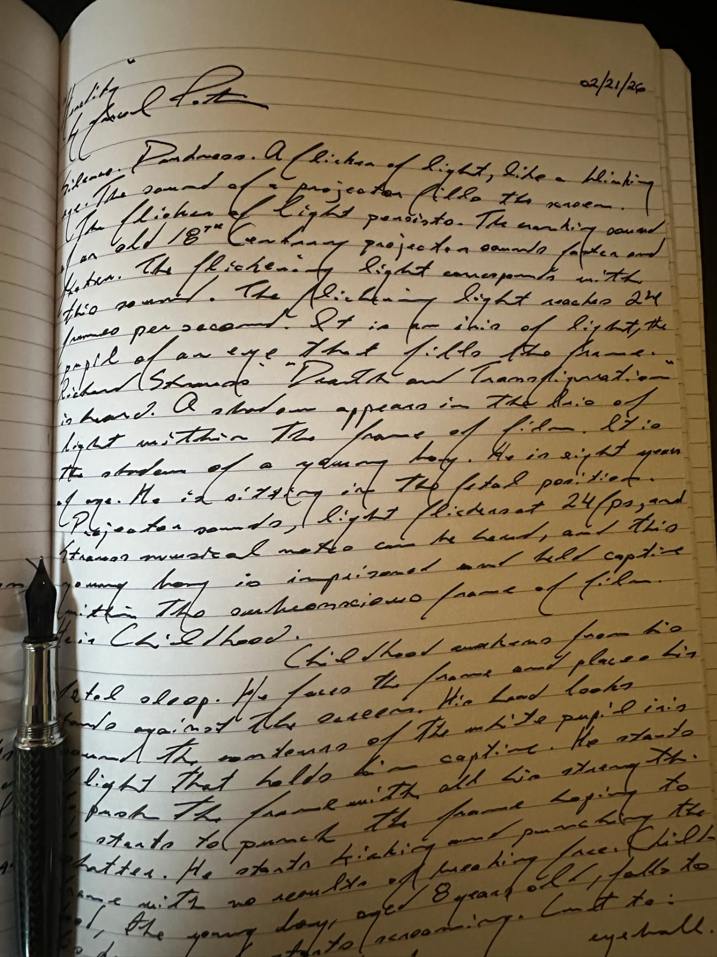 Notebook page in Marcel Potvin's fountain-pen handwriting, dated 02/21/26 and headed Heredity, opening 'Silence. Darkness. A flicker of light, like a blinking eye. The sound of a projector fills the screen', with a black fountain pen resting on the left