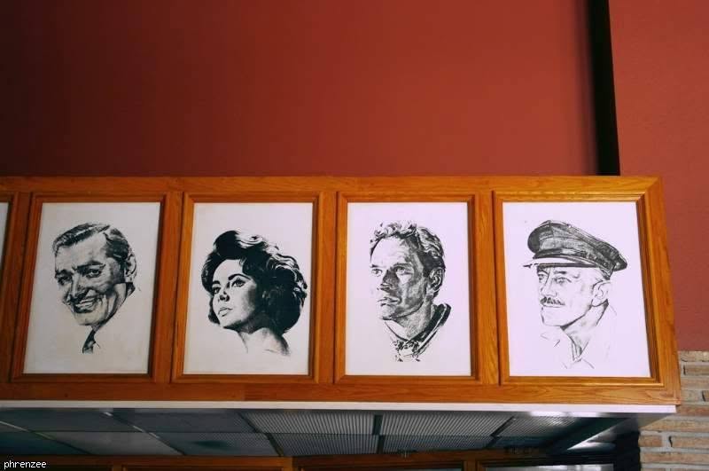 Close-up of four framed black-and-white pencil drawings of classic Hollywood stars in a wooden frame above the concession stand, against a deep red wall.