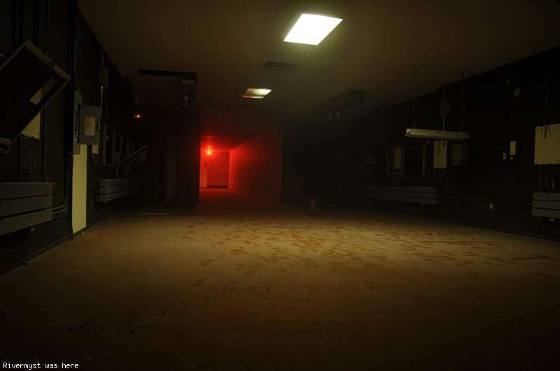 A long dark back hallway lit by a few ceiling lights, glowing red at the far end from an exit sign.