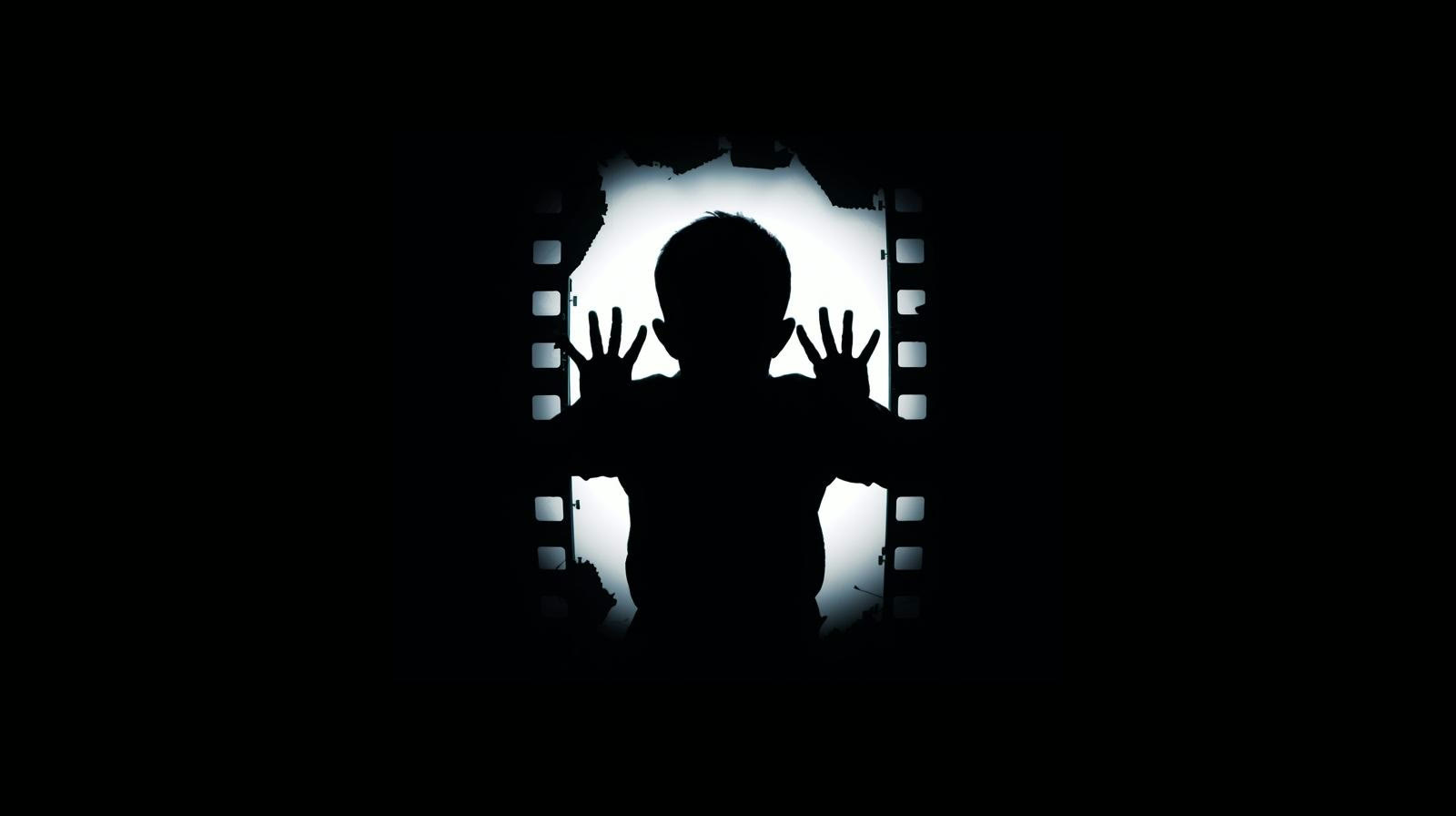 A child's silhouette presses both hands against a window of light framed by film perforations