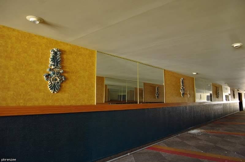 The long hallway to the auditoriums, lined with gold walls, tall mirrors and ornate blue-and-cream wall sculptures above a dark blue lower wall, with a patterned carpet striped in red and yellow.