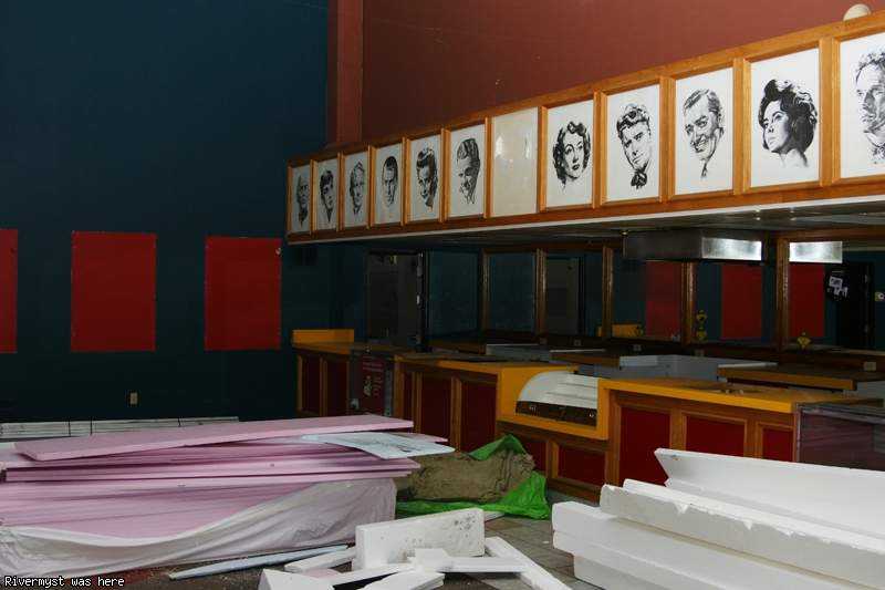 The empty concession stand with red and yellow counters, topped by a row of framed black-and-white drawings of classic movie stars, with foam boards piled on the floor.