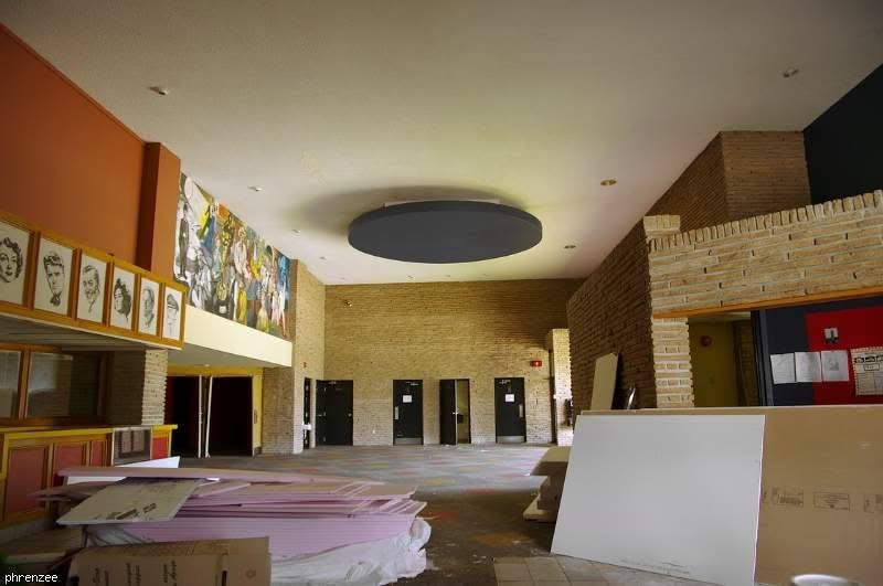 The wide main lobby with tall brick walls, a round dark disc hanging from the ceiling, the painted movie-star mural high on the left above the concession stand's framed star portraits, and a row of black doors at the far end.